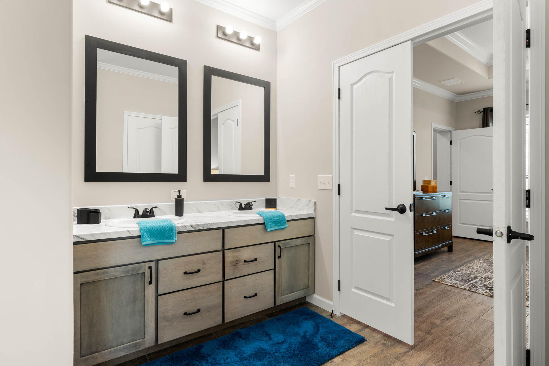 Modern bathroom with dual sinks, wooden vanity, and two large mirrors. Bright lighting, teal towels, and a plush blue mat add a fresh feel. Doorway leads to adjacent room.