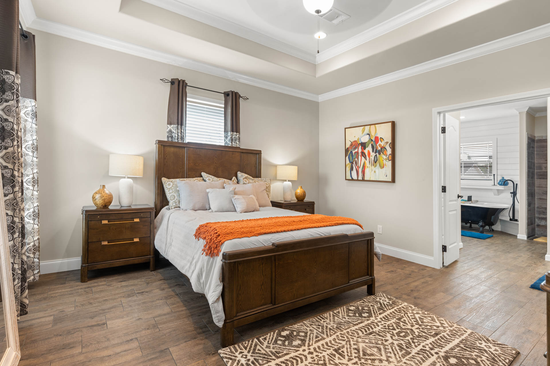 Cozy bedroom with a wooden bed, white and orange bedding, two bedside tables with lamps, abstract wall art, patterned rug, and attached bathroom.