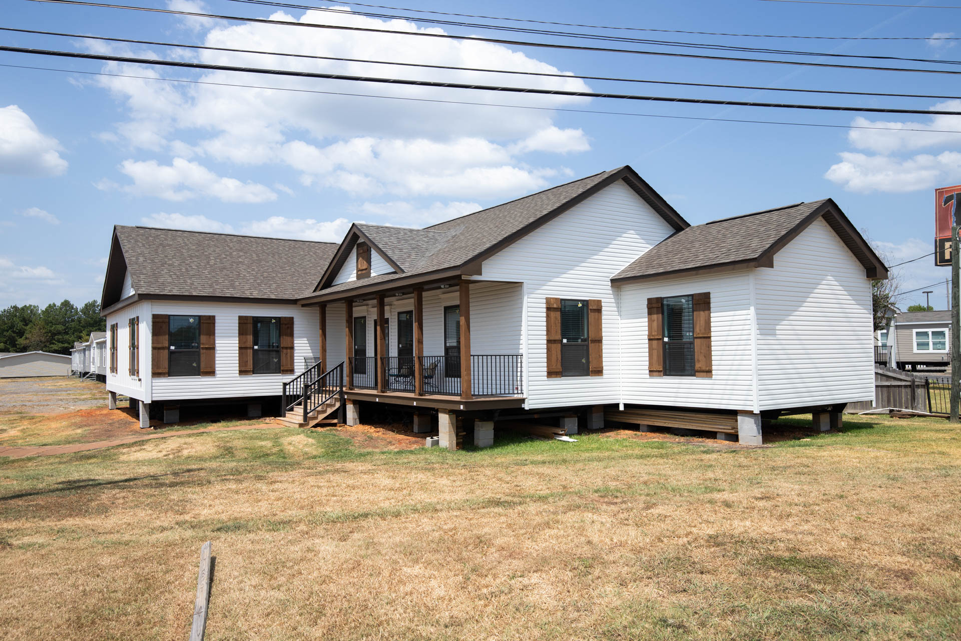 White modular house with a dark shingled roof, elevated on stilts, sits on a dry grassy lot under a blue sky with clouds. Simple and modern.