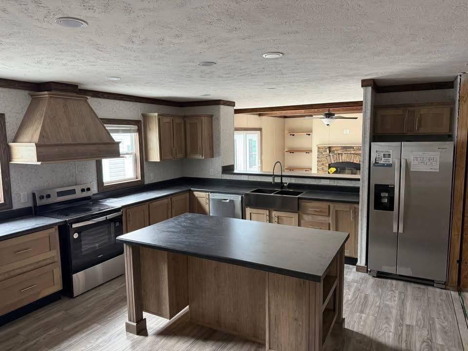 Spacious kitchen with wood cabinets and a center island. Stainless steel appliances include a fridge and oven. A sink overlooks a bright, cozy living room.