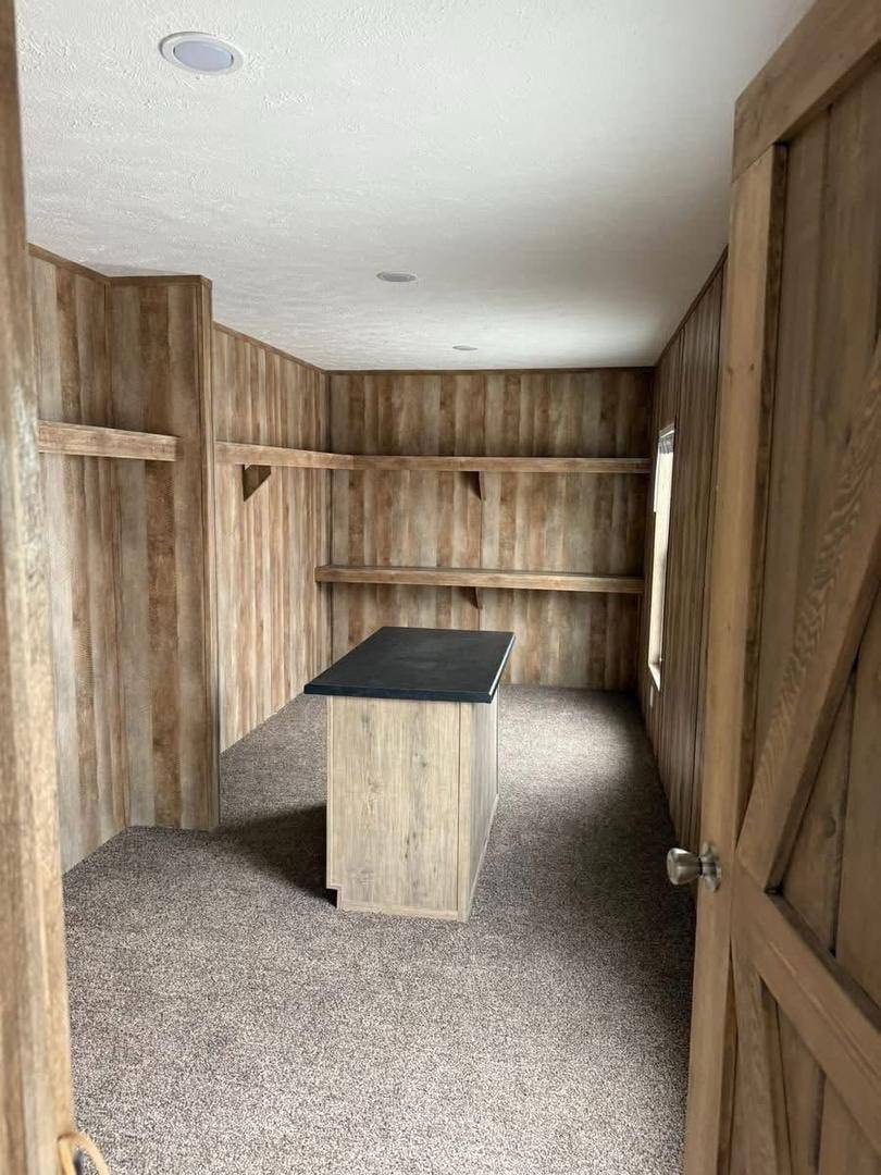 A small, rustic room with wood-paneled walls and shelves, featuring a central island with a dark countertop. The carpet is brown, creating a cozy feel.