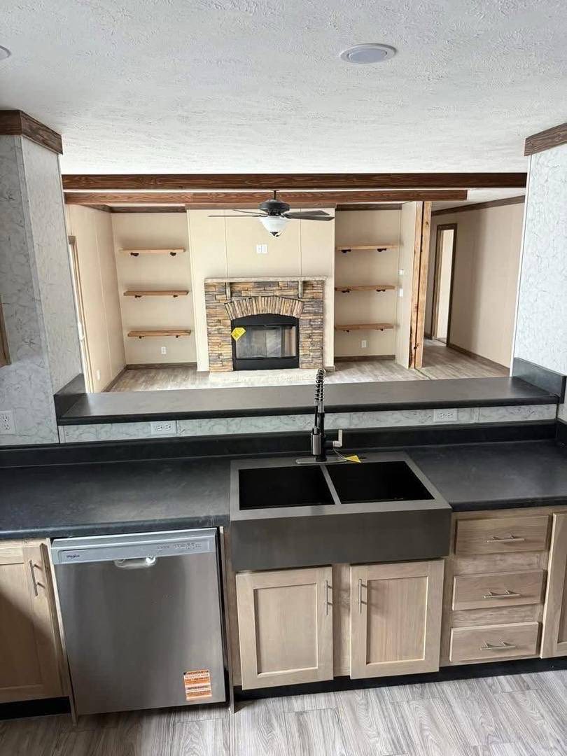 Spacious kitchen with light wood cabinets, dark countertops, and stainless steel dishwasher. The adjacent living room features a stone fireplace and wooden shelves.