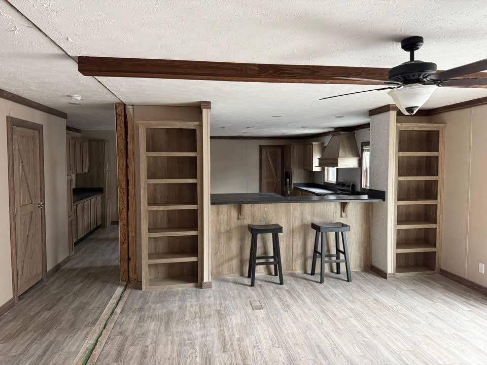 Spacious modern kitchen with light wood floors, two open shelves flanking a breakfast bar with two stools. Ceiling fan and neutral tones create a cozy feel.