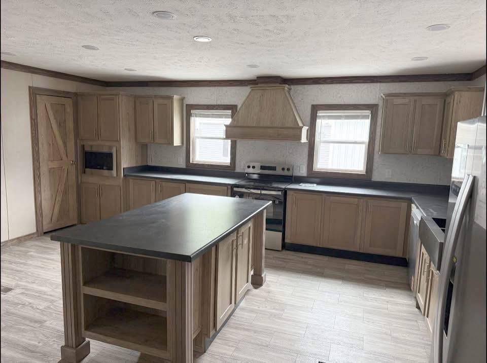 Spacious kitchen with light wood cabinetry and dark countertops. Central island, stainless steel appliances, vent hood, and large windows enhance brightness.