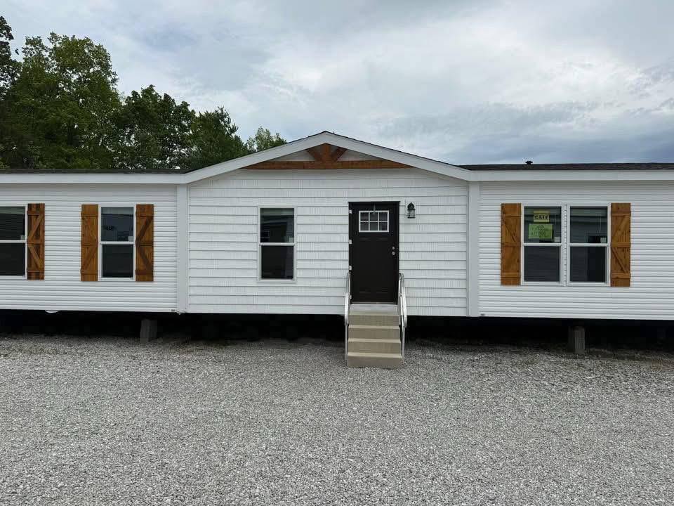 A white manufactured home with wooden shutters and a black door stands on a gravel lot. Steps lead to the entrance, framed by trees and a cloudy sky.