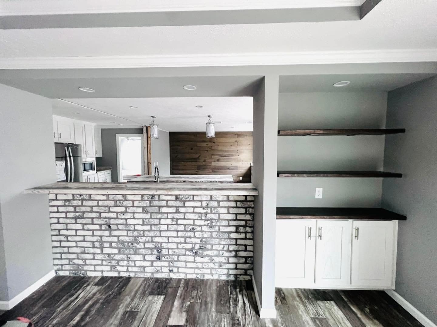 Modern kitchen interior with light gray walls and wood flooring. Features a brick bar, white cabinets, dark wooden shelves, and pendant lights, creating a cozy ambiance.