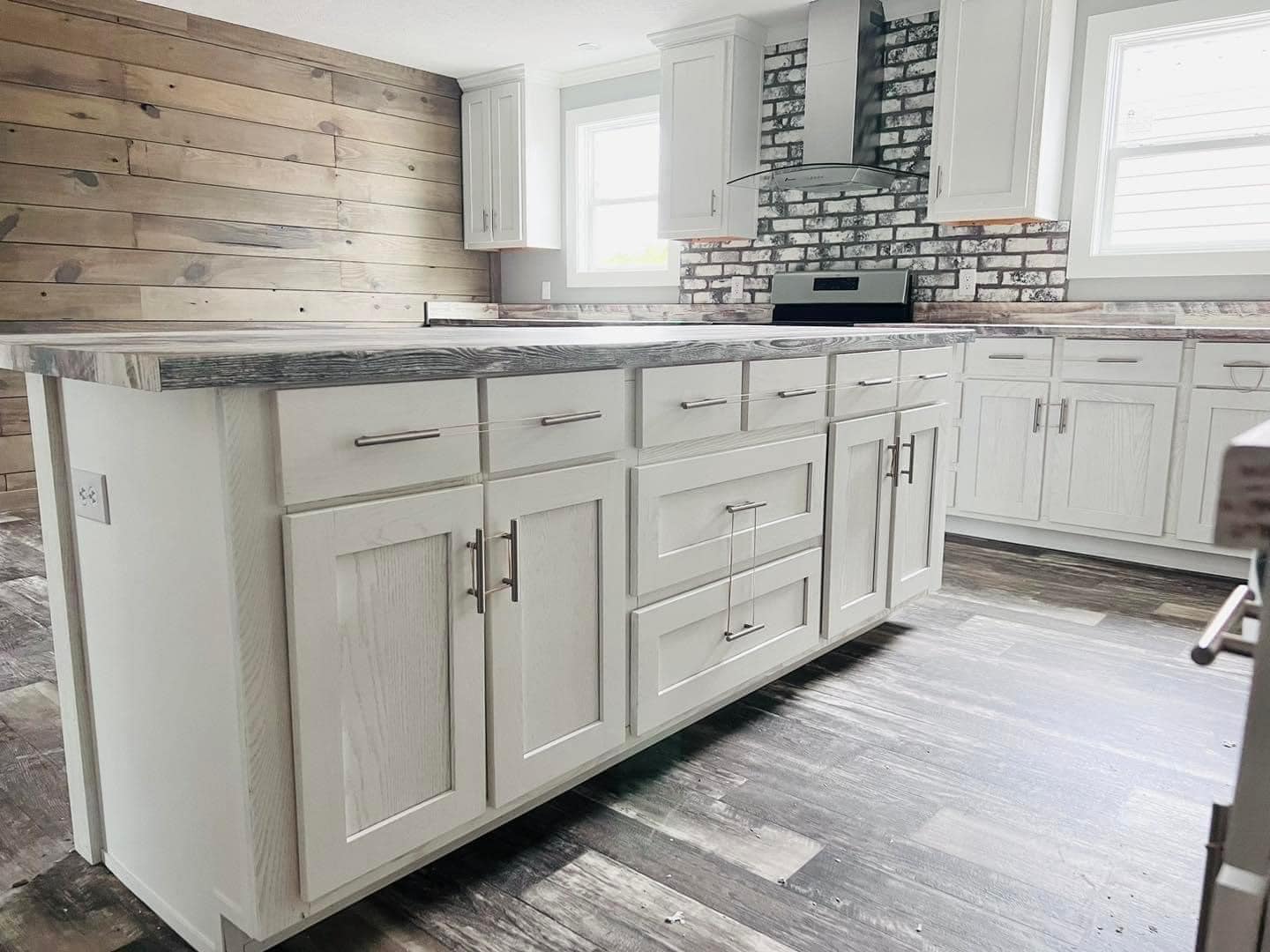 A modern kitchen with white cabinets, a gray wood island, and stainless steel handles. Brick backsplash and wood panel accent wall create a rustic feel.