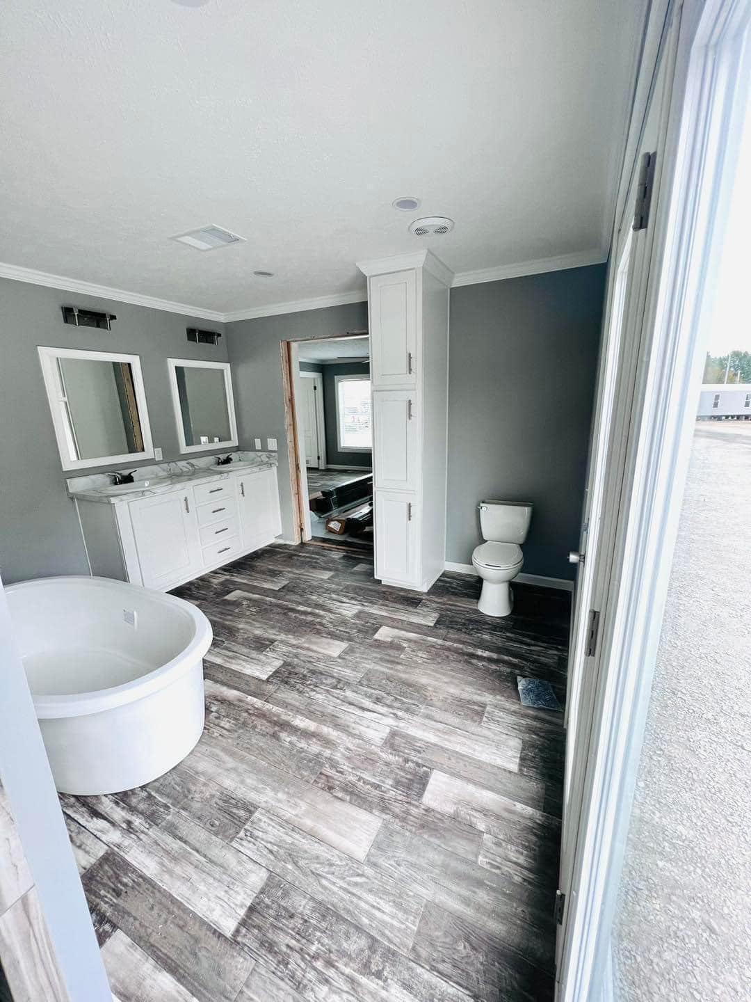 Modern bathroom with gray walls, wood-look floor, white vanity, dual mirrors, deep round tub, and toilet, conveying a clean, minimalist feel.