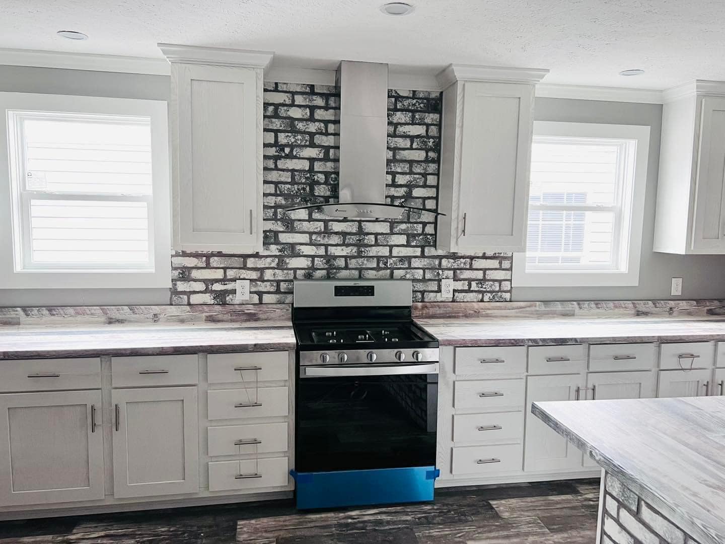 Modern kitchen with white cabinets, a stainless steel stove, and a brick backsplash between two windows. The atmosphere is bright and clean.