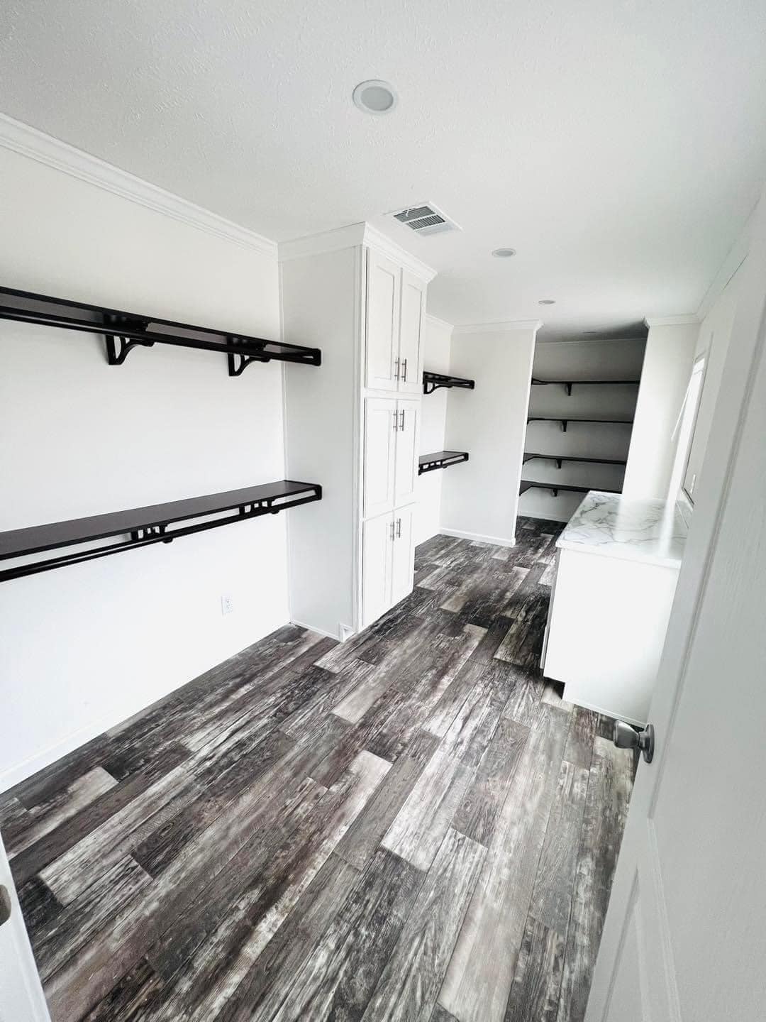 Narrow storage room with white walls, dark wood floor, and multiple black shelves. White cabinets and a window add brightness, creating an organized, airy feel.