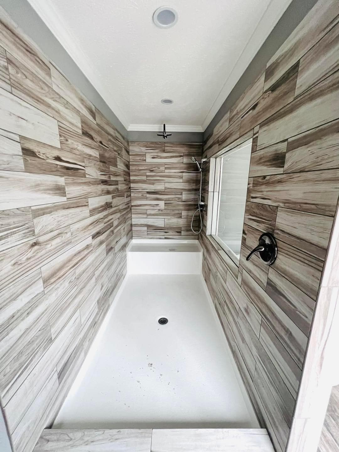 Narrow bathroom with a white tub and shower. Walls are tiled with wood-like patterns, creating a rustic feel. A window on the right brings in natural light.