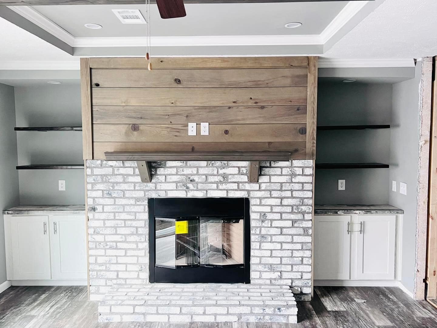 Living room with a rustic vibe featuring a whitewashed brick fireplace, wooden mantel, gray walls, floating shelves, and white cabinets on both sides.