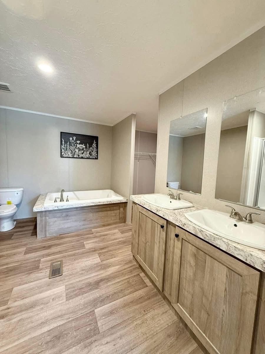 Spacious bathroom with wood flooring, featuring a double sink vanity, a large bathtub, toilet, and floral wall art. Bright, clean, and serene atmosphere.