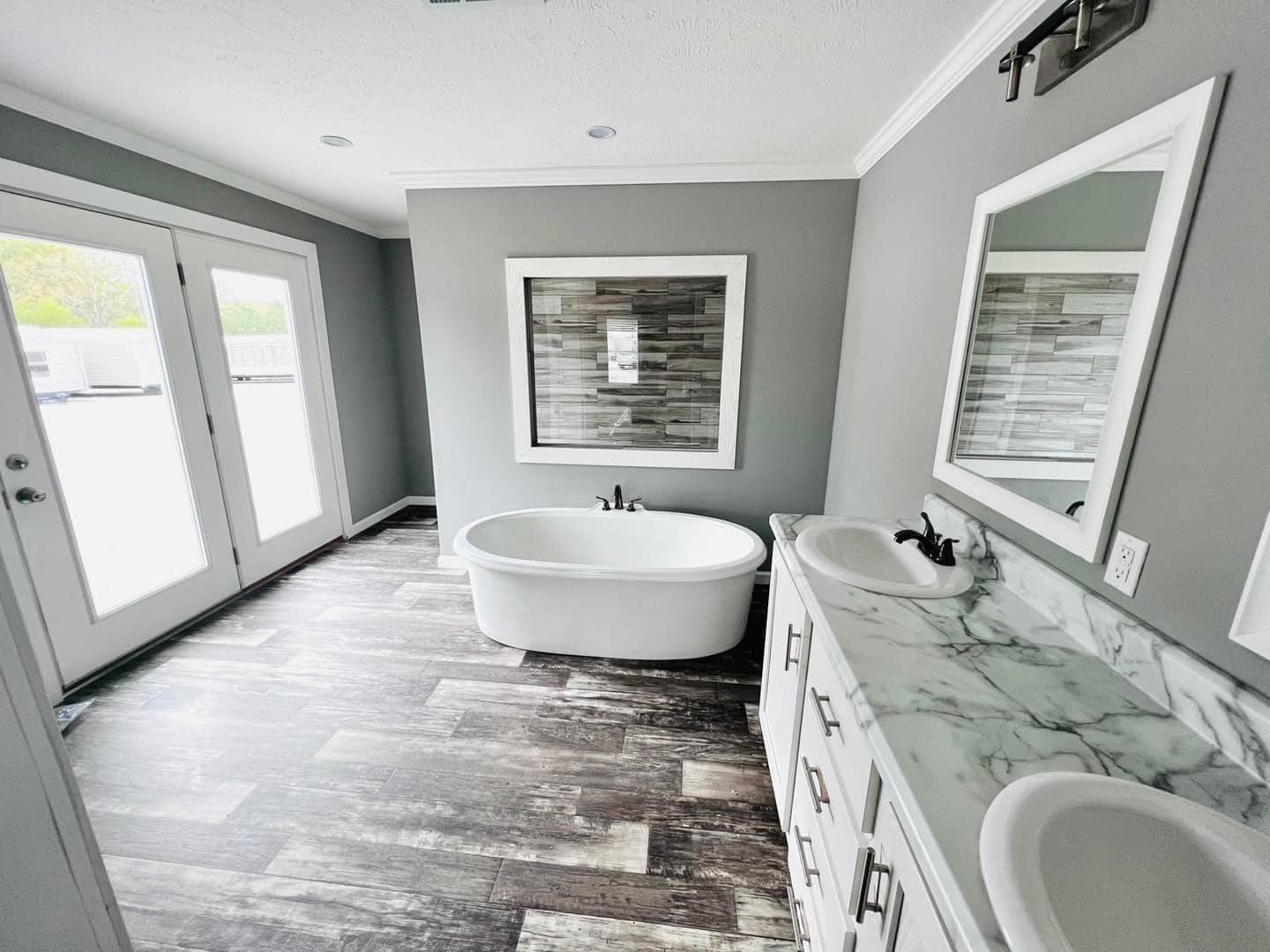 Bright modern bathroom with gray walls and wood-patterned floor. Features a freestanding bathtub, marble countertop, dual sinks, and French doors.