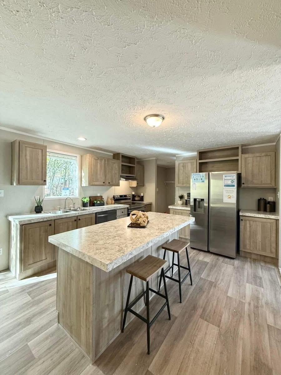 Spacious kitchen with wooden cabinets and flooring, featuring a large island with two stools, stainless steel fridge, and bright natural light. Cozy and inviting.