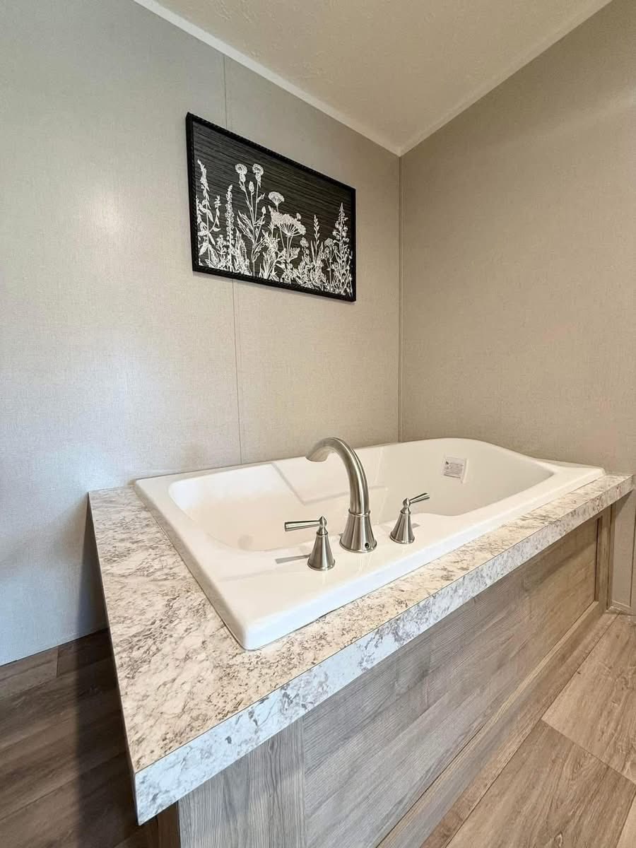 Modern bathroom with a white rectangular bathtub featuring silver faucets, set in a wood-paneled enclosure. A botanical print adorns the beige wall.