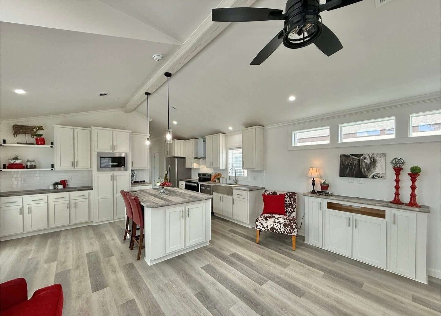 Modern kitchen with white cabinets, gray countertops, and island seating. Red accents throughout, wood flooring, ceiling fan, and soft lighting create a cozy vibe.