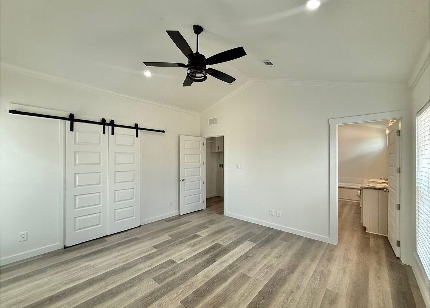 A spacious, modern room with light wood flooring and white walls. There's a ceiling fan, barn-style closet doors, and an open door leading to a bright adjoining room.