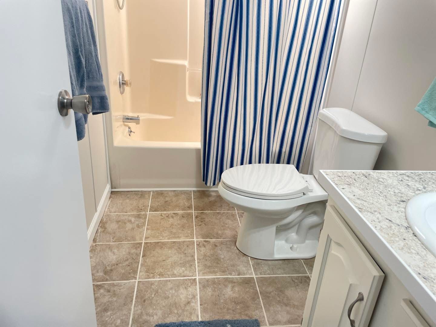 A clean bathroom with beige tiled floor, white toilet, and a bathtub with a blue and white striped shower curtain. Towels hang on the door and wall.