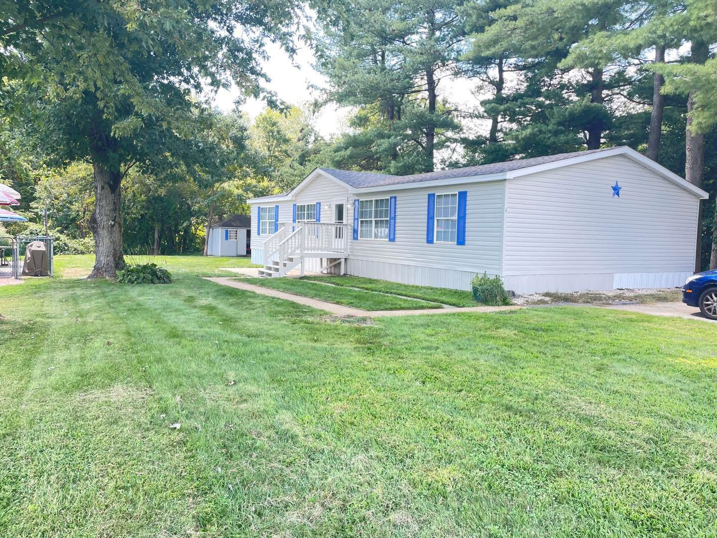 Single-story white house with blue shutters and a blue star on the side. Surrounded by green grass and tall trees, creating a peaceful atmosphere.