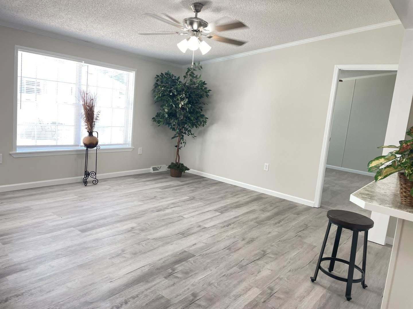 Bright, spacious living room with light wood flooring, beige walls, and a large window. Features a ceiling fan, potted plants, and a bar stool. Cozy and modern atmosphere.
