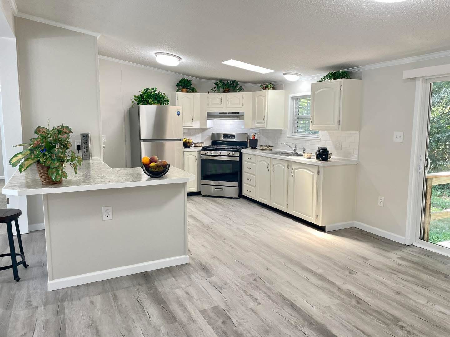 A bright, modern kitchen with white cabinets, stainless steel appliances, and light wood flooring. Plants and a fruit bowl add a fresh touch.