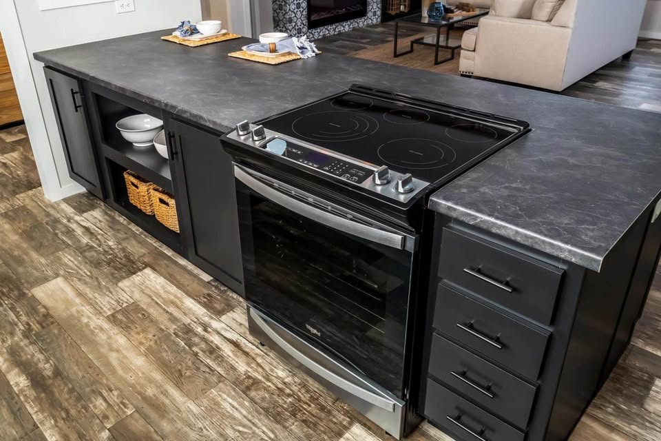 Modern kitchen with a sleek, black countertop featuring an electric stove. Open shelving and drawers below, wooden flooring, and a cozy living area in the background.