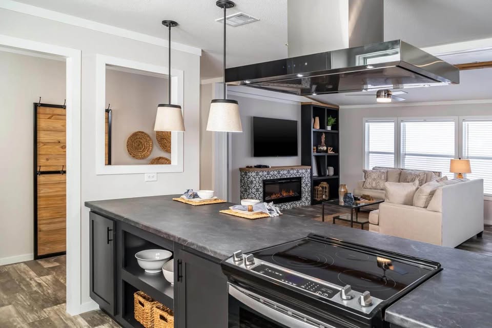 Modern open-concept kitchen and living room with sleek black countertops, pendant lights, and cozy beige sofa. A TV and fireplace enhance the inviting space.