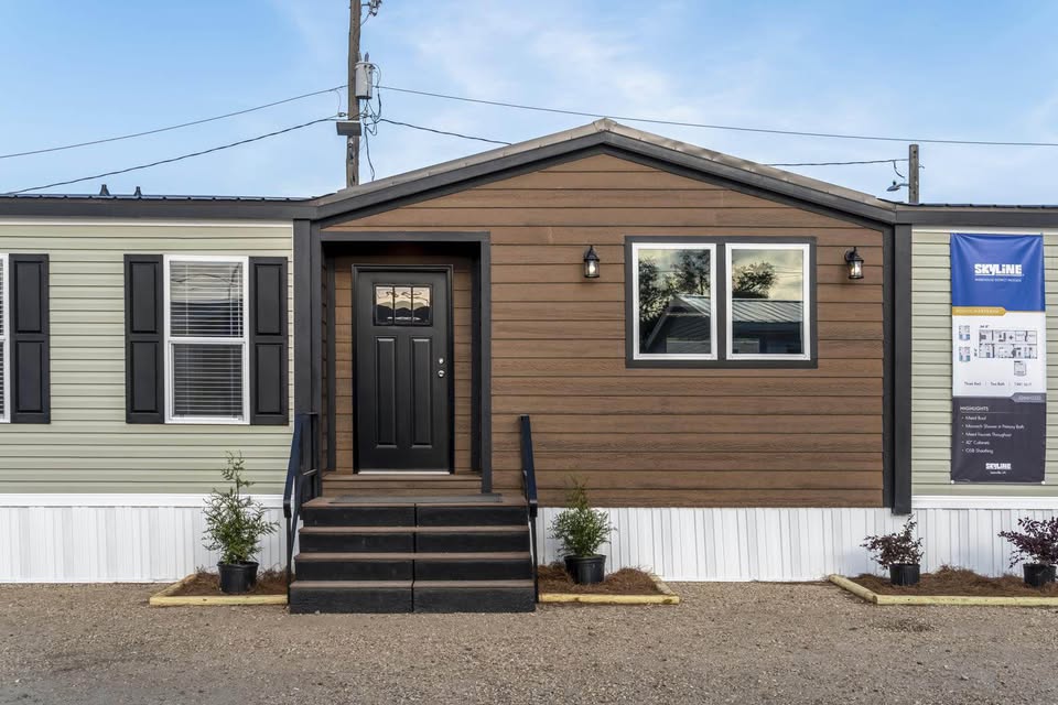 Single-story modular home with a brown central facade, black door, and steps. Flanking light-gray sections have windows and a banner. Small plants line the front.