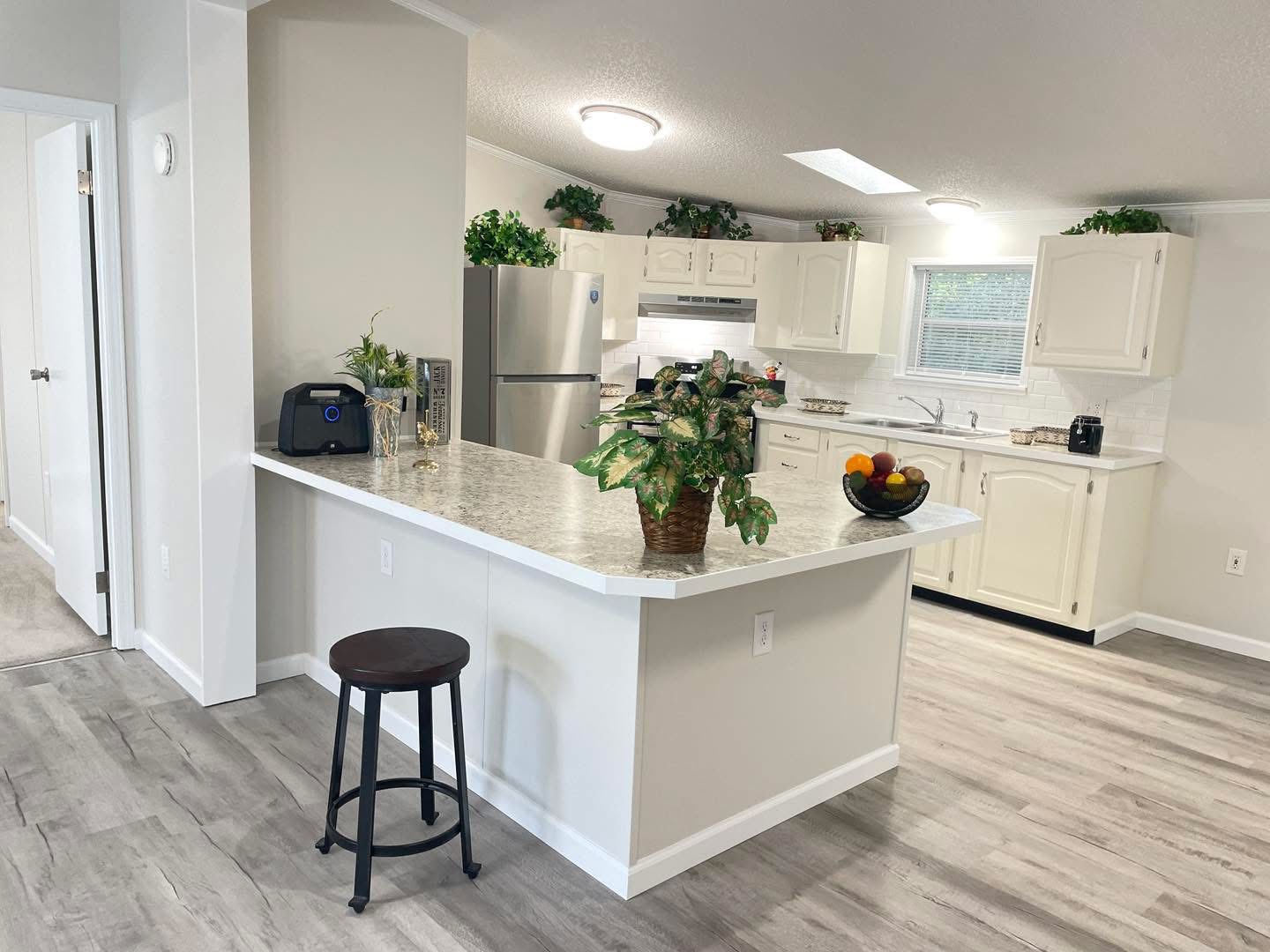 Bright kitchen with white cabinets, gray countertops, and light wood floors. Includes stainless steel appliances, potted plants, and a fruit bowl on the island.
