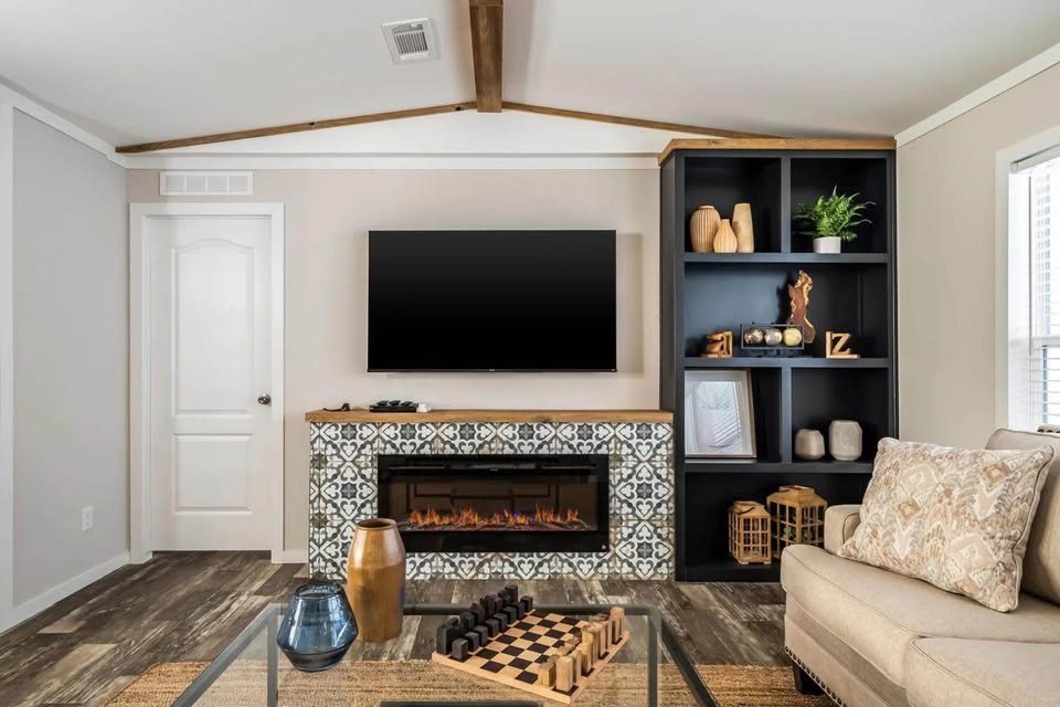 Modern living room with a patterned tile fireplace below a wall-mounted TV. A cozy beige couch is on the right, with a glass coffee table holding vases and a chessboard in the foreground. A black bookshelf with decor and a potted plant adds contrast. Warm, inviting atmosphere.