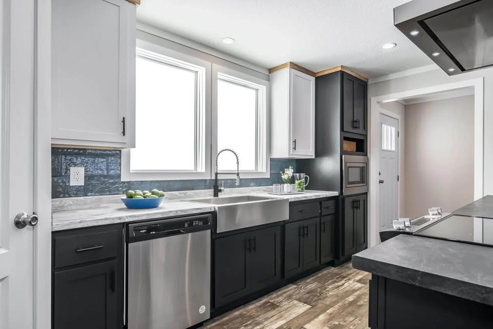 Modern kitchen with black and white cabinets, stainless steel appliances, and a farmhouse sink. Wooden flooring and bright natural light create a sleek, inviting atmosphere.