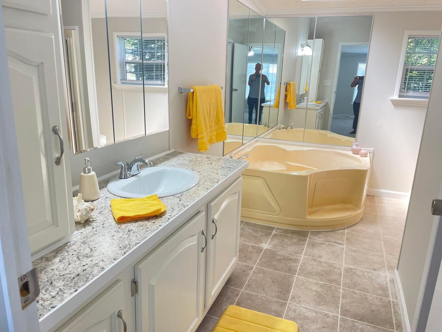A cozy bathroom featuring a beige tub, white cabinets with granite countertops, and yellow towels. Mirrors enhance space, creating a welcoming atmosphere.