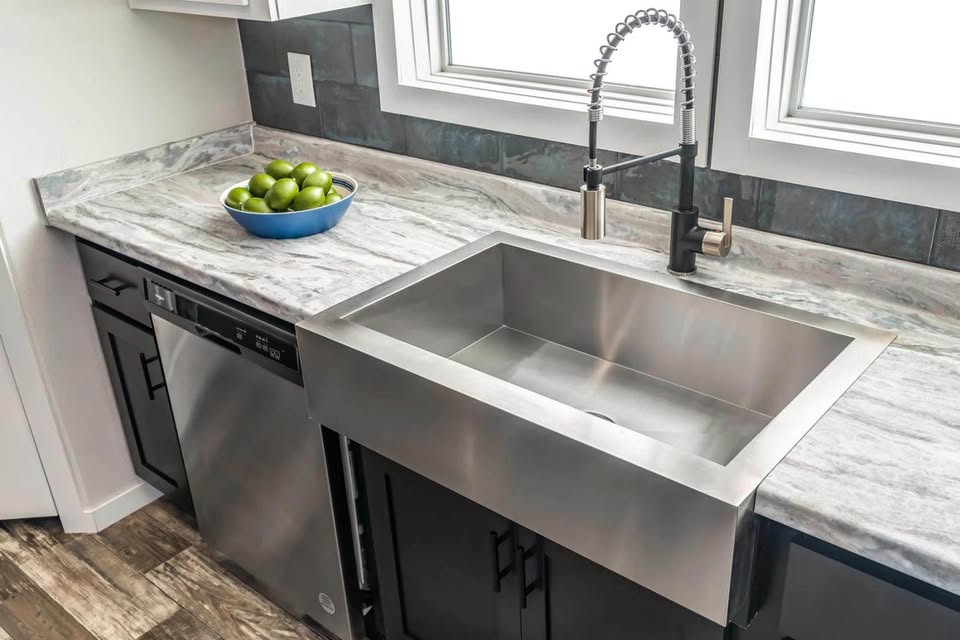 Modern kitchen with stainless steel sink, sleek faucet, and marble countertop. Blue bowl of green apples adds a pop of color on the left. Bright, clean feel.