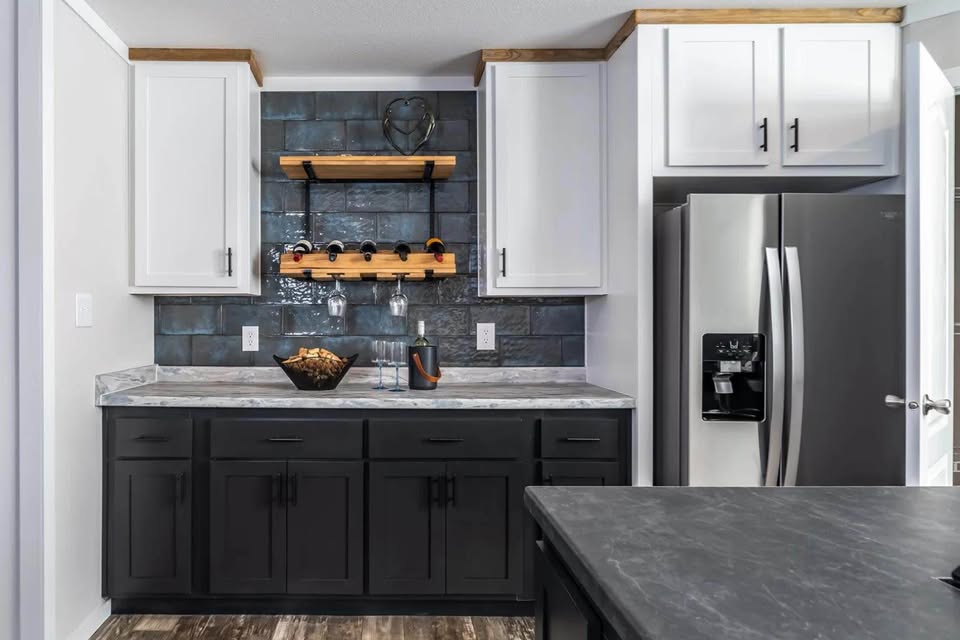 Modern kitchen with gray and white cabinets, black backsplash, and marble countertop. Features a wooden wine rack, two glasses, and a stainless steel fridge.