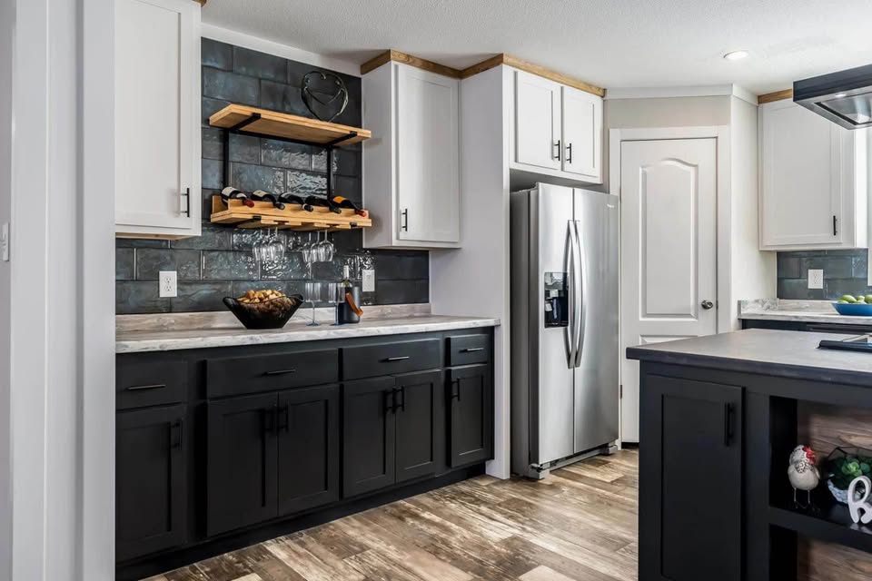 Modern kitchen featuring dark lower cabinets and white upper cabinets with wooden trim. A stainless steel fridge, open shelves holding wine bottles, and a bowl of fruit add to the sleek, contemporary feel.