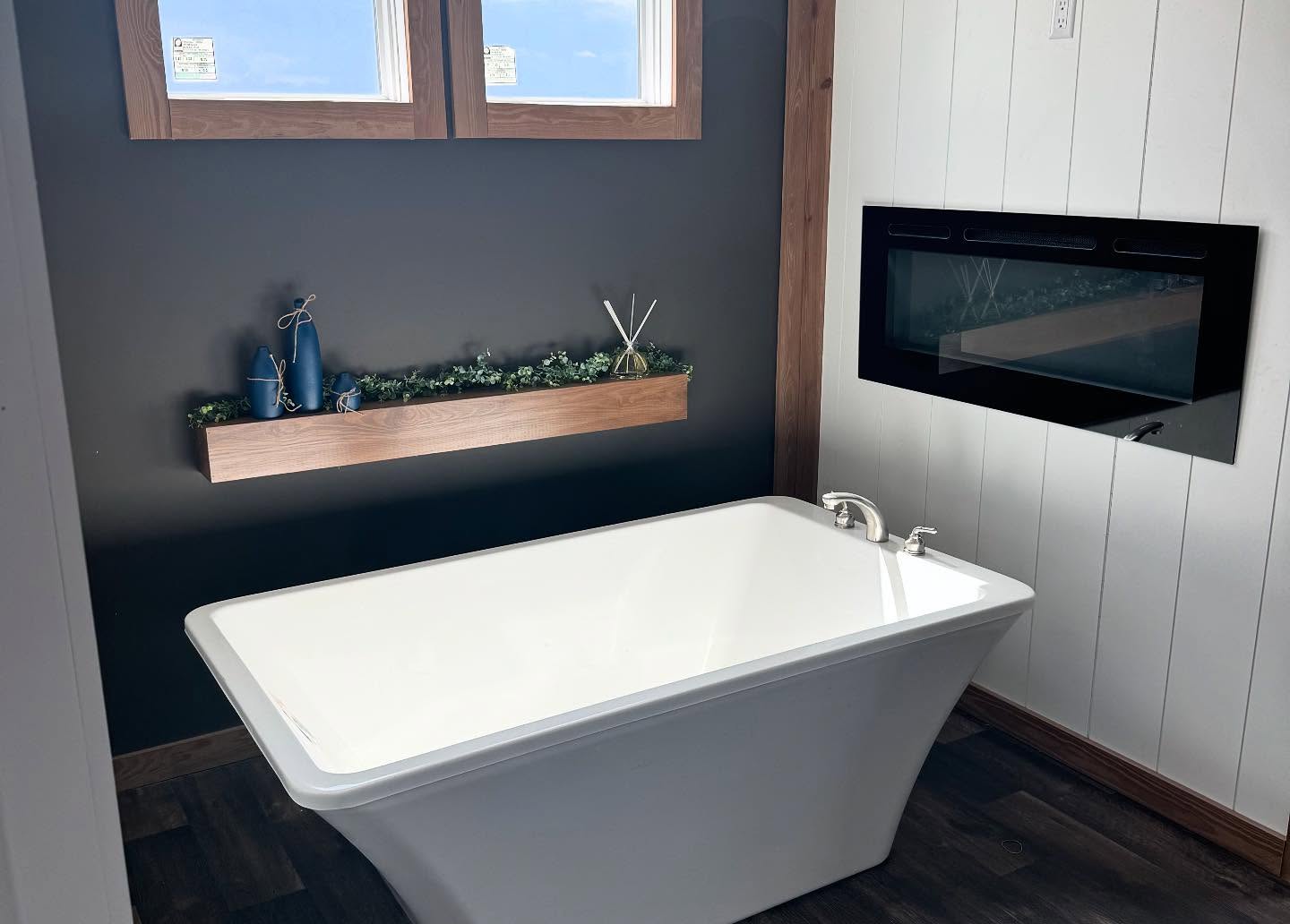 Modern bathroom with a white freestanding tub, black accent wall, wooden shelf with greenery, two blue candles, and a sleek electric fireplace. Cozy ambiance.