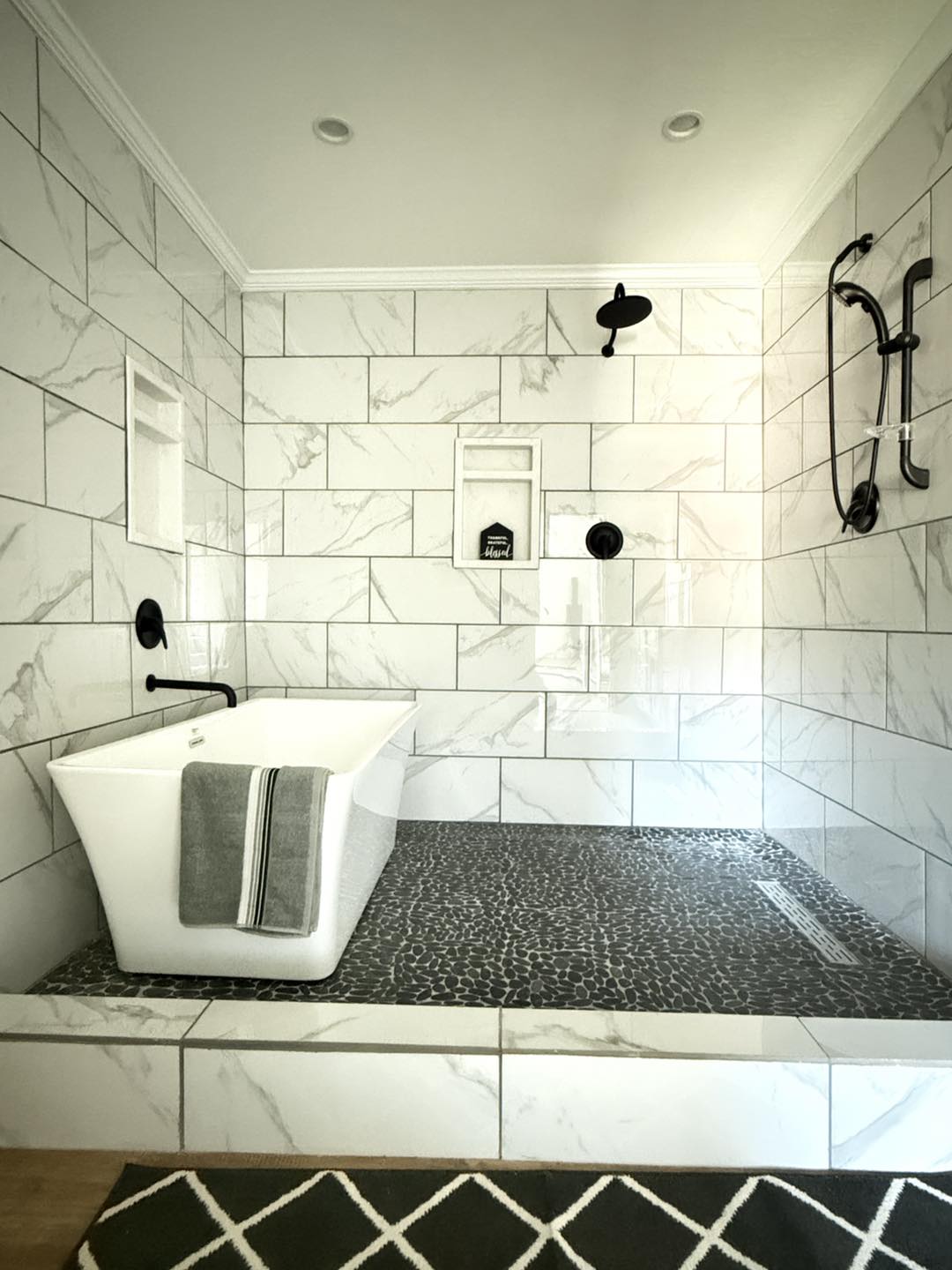 Modern bathroom with a sleek white bathtub, gray towel, and black fixtures. Marble tile walls create a calm, elegant atmosphere. Geometric floor mat.