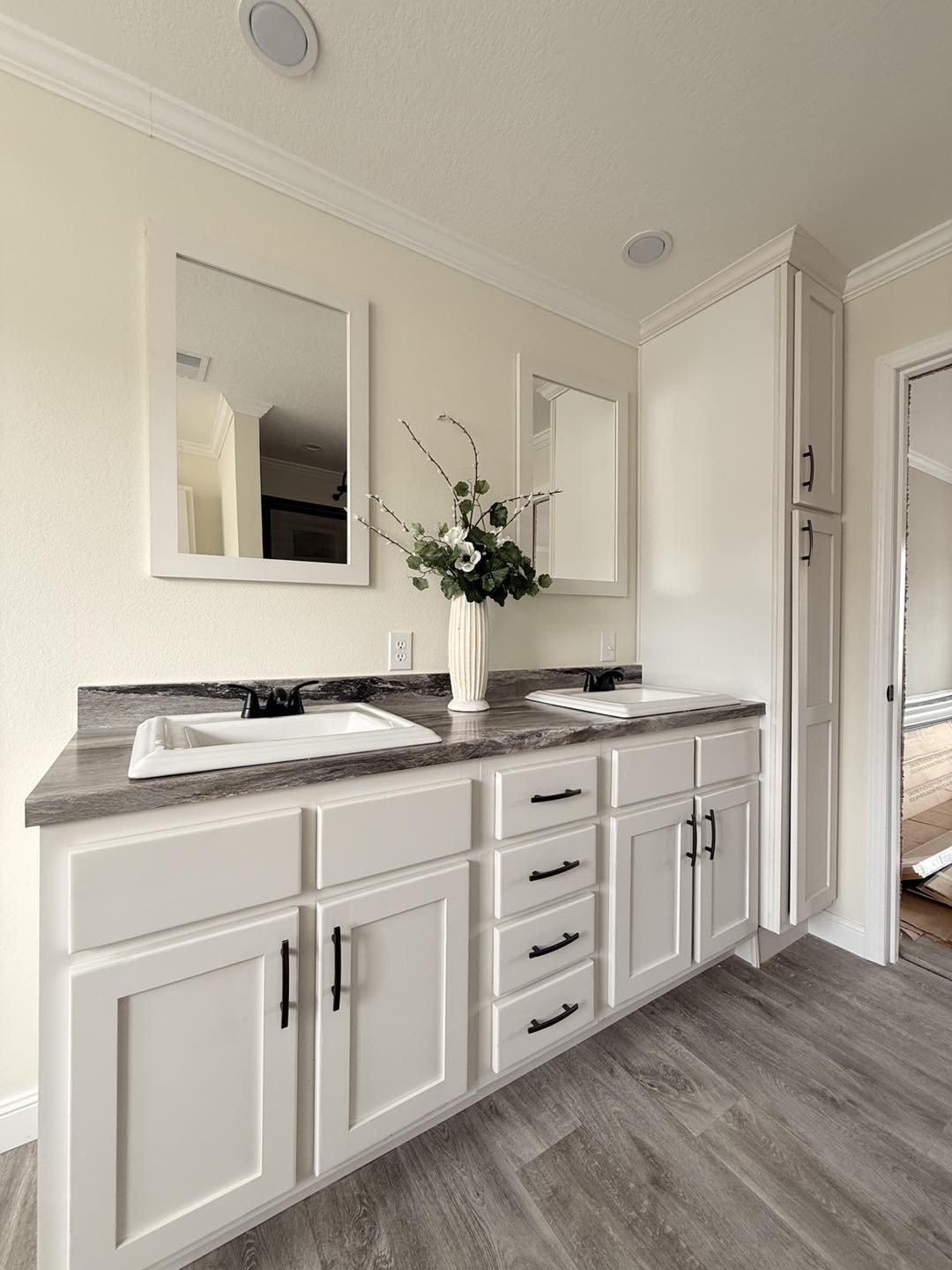 Modern bathroom with dual sinks on a marble countertop, white cabinets, and a vase with flowers. Light walls and wooden flooring create a serene feel.