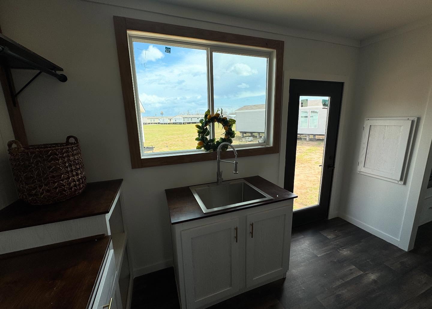 A small kitchen with dark wood counters, a stainless steel sink, and a large window adorned with a sunflower wreath. Natural light and a serene outdoor scene are visible.