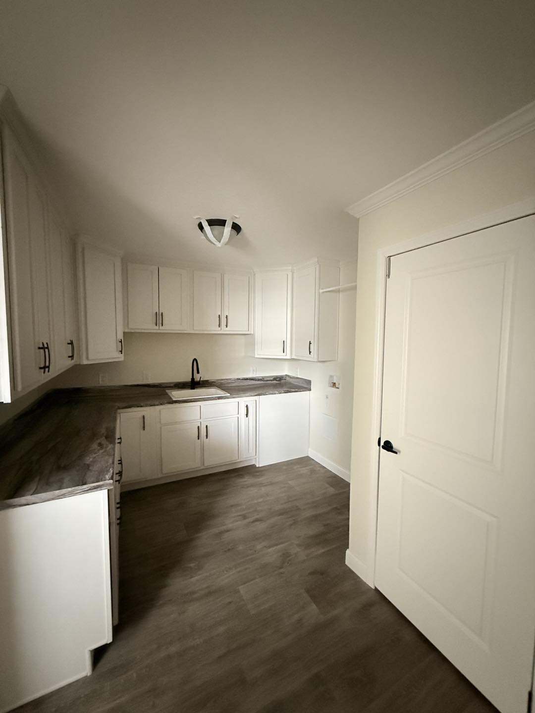 A small kitchen with white cabinets, dark marble countertops, and stainless steel handles. The wooden floor and neutral tones create a cozy atmosphere.