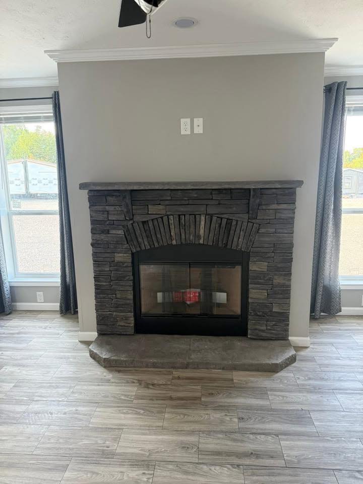 Stone fireplace with a modern design, centered between large windows. Gray walls and wood flooring create a cozy yet contemporary atmosphere.