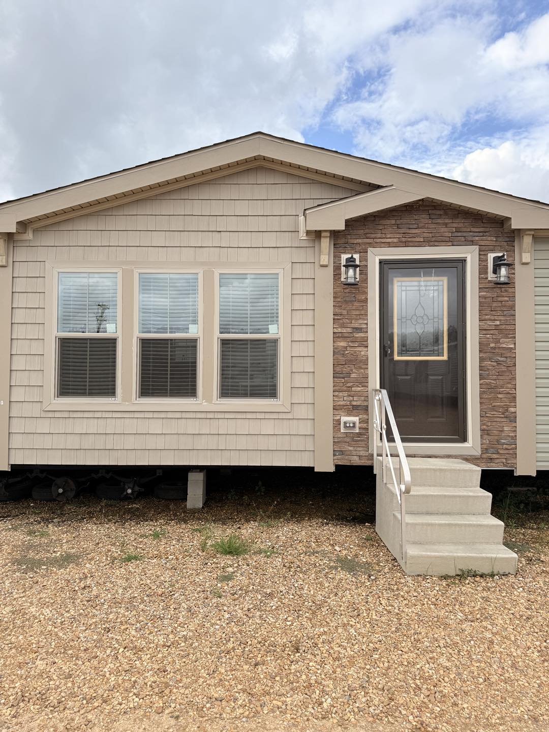 A beige, single-story mobile home with a stone accent near the entrance, three large windows, and a staircase leading to the front door under a cloudy sky.