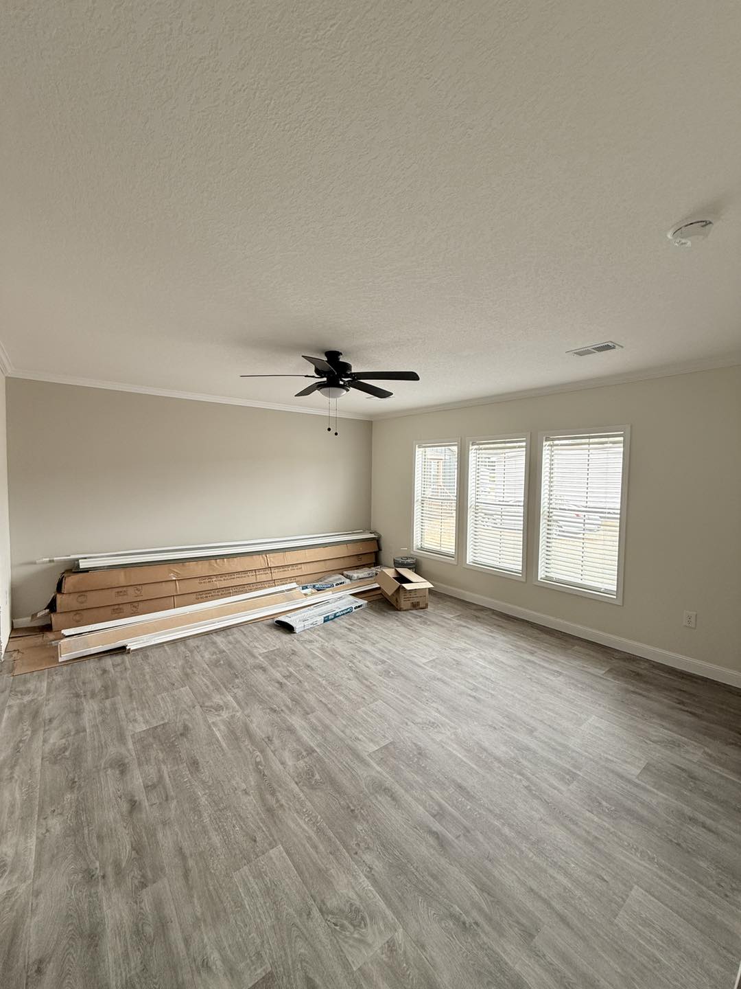 Spacious empty room with light wood flooring, neutral walls, and a ceiling fan. Three large windows let in natural light; construction materials lie on the floor.