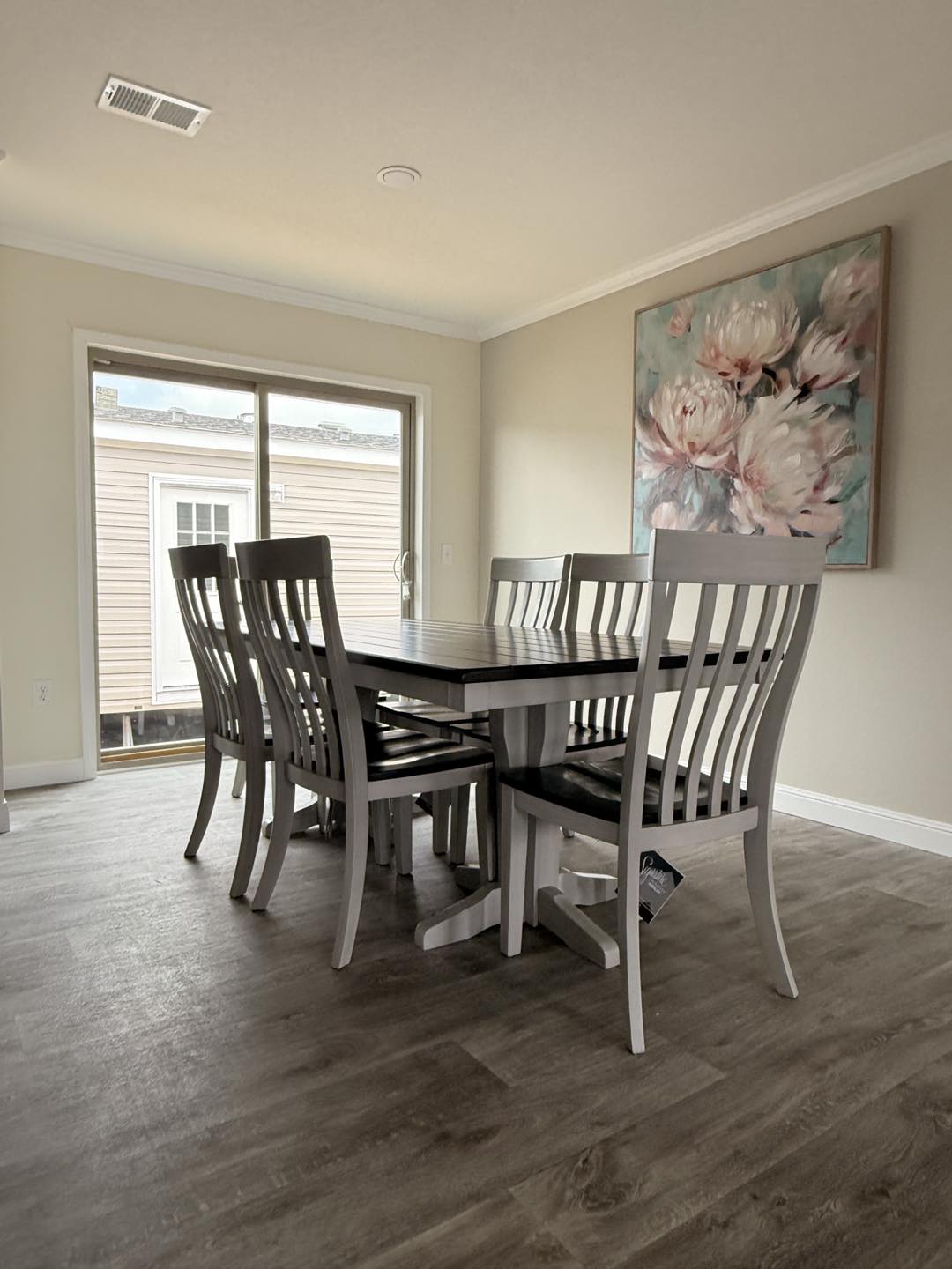 Dining room with six gray wooden chairs around a dark wooden table. A large floral painting hangs on the light wall, with soft natural light streaming in.