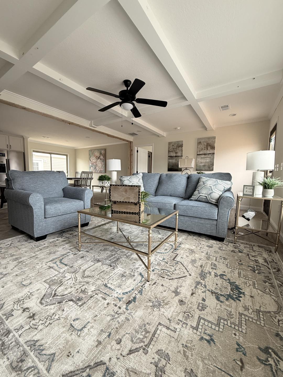 Spacious living room with gray sofas, patterned cushions, and glass coffee table atop a neutral, patterned rug. White walls, ceiling beams, and fan. Cozy ambiance.