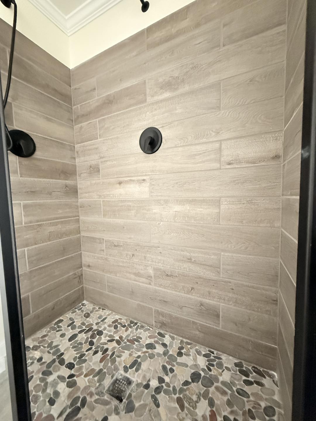 A modern shower interior featuring light wood-like tiles and a pebble stone floor. The dark showerhead adds contrast, creating a calm, earthy ambiance.