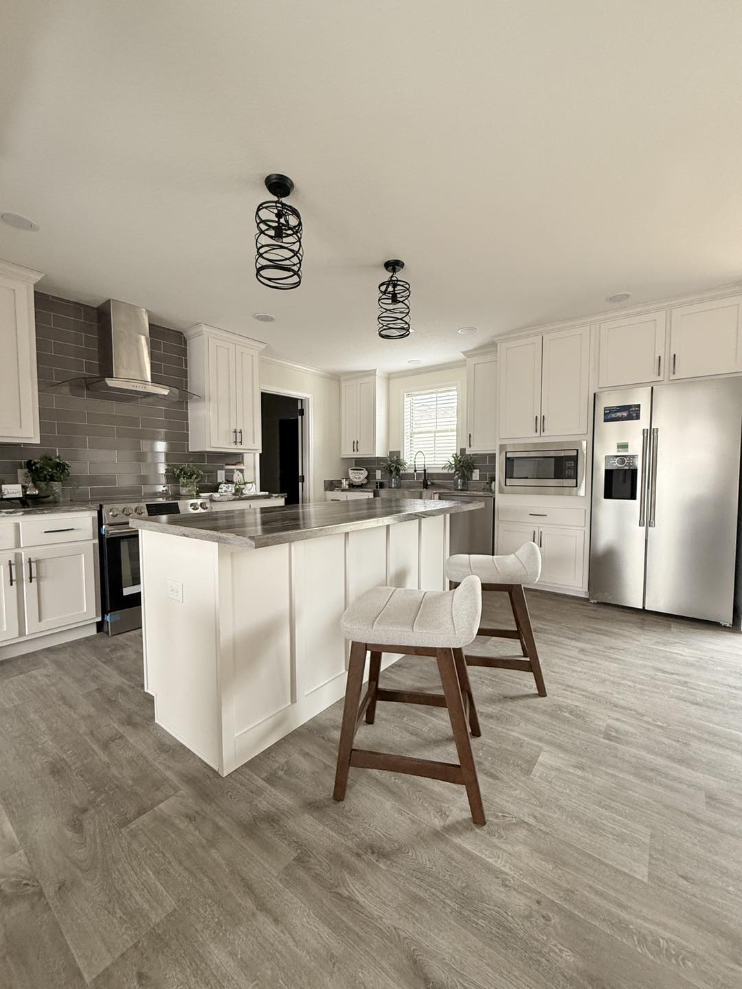 Modern kitchen with white cabinets, gray backsplash, and stainless steel appliances. Central island features two cushioned stools and pendant lights.