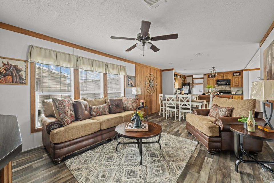 Spacious living room with two brown sofas, a patterned rug, and a round coffee table. Ceiling fan above, dining area with white chairs in background. Cozy and inviting ambiance.