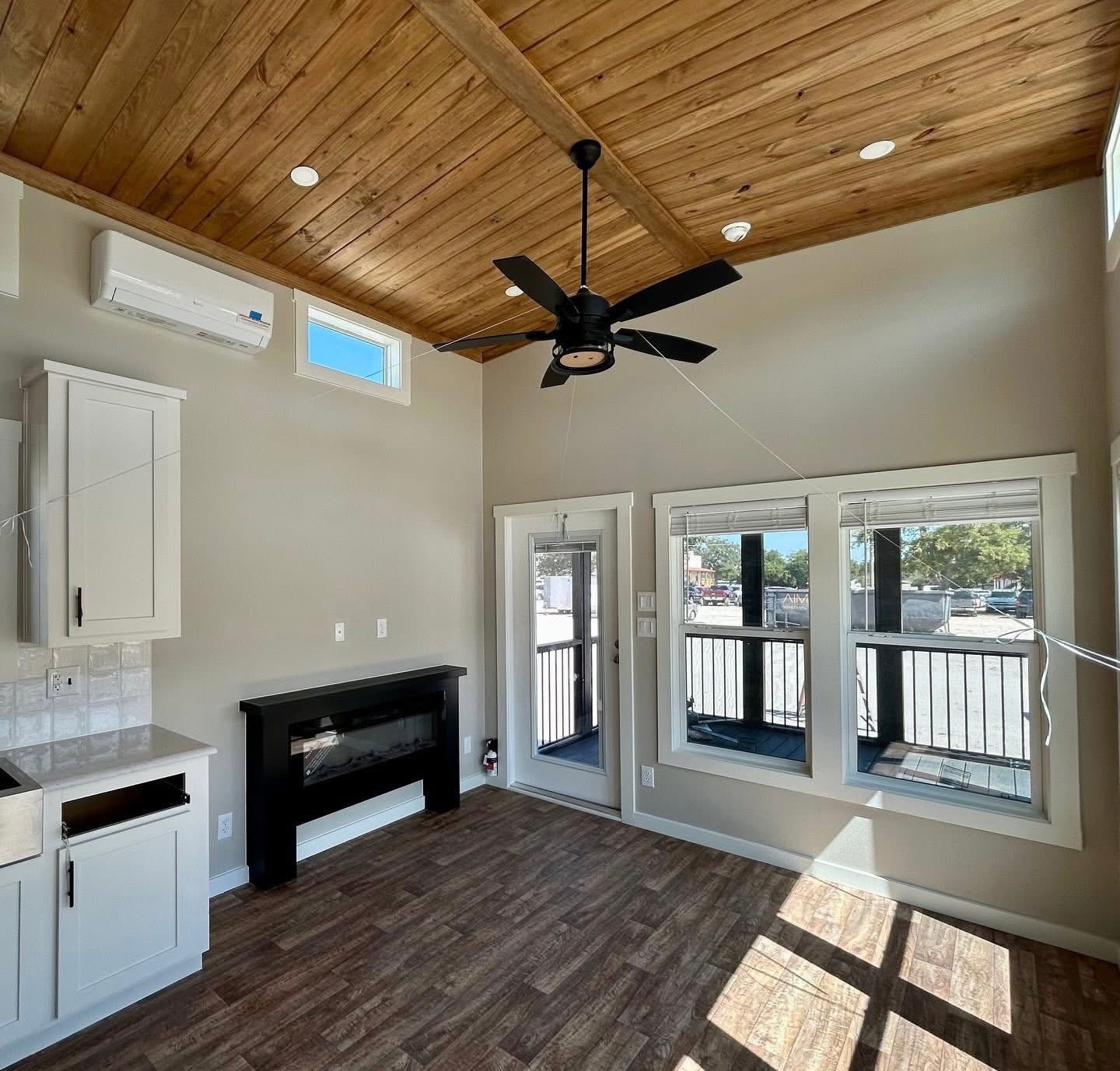Cozy room with wooden vaulted ceiling, fan, and large windows. Features include white cabinets, an electric fireplace, and rich wood flooring. Bright and airy atmosphere.